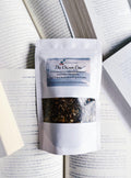 A packaged product labeled 'The Chosen One' from The Sunny Teacup, featuring a mix of dried herbs visible through the packaging window, placed on top of an open book.